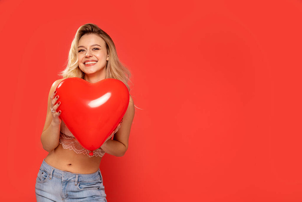 Happy Caucasian blonde beautiful lady on Valentines Day, smiling and enjoying romantic holiday. Girl holding red heart shaped balloon. A lot of copy space - Fotografie, Obrázek