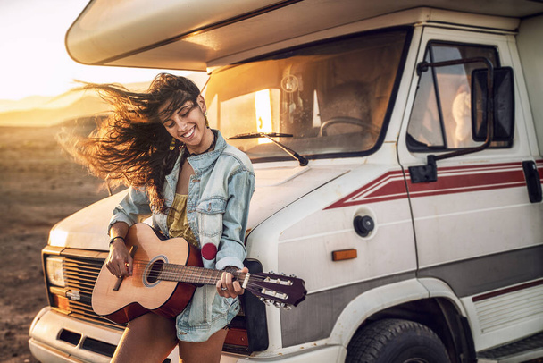 Hipster beautiful woman playing guitar with big smile on her face. Nomadic and camper lifestyle concept. Warm sunset light.  - Foto, afbeelding