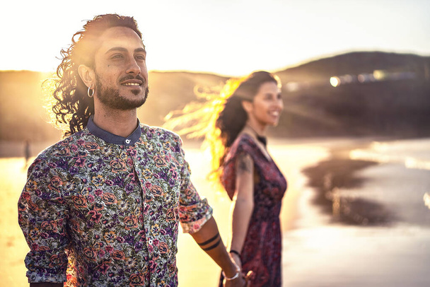 Attractive italian couple holding hands and looking together at the same direction. Romantic date at sunset on the beach .Real people emotions and love concept. Focus on the man. - Foto, Bild