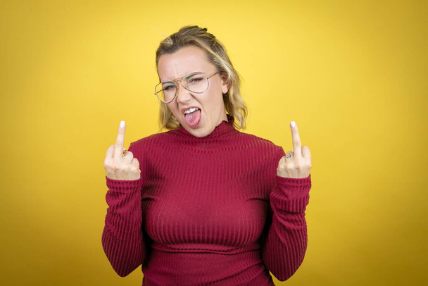 Young caucasian woman wearing casual red t-shirt over yellow background showing middle finger doing fuck you bad expression, provocation and rude attitude. screaming excited - Fotó, kép