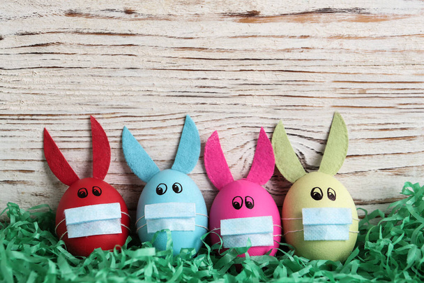 Dyed eggs with bunny ears in protective masks on wooden background, flat lay and space for text. Easter holiday during COVID-19 quarantine - Photo, Image
