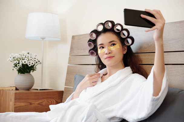 Bella giovane donna asiatica con rulli per capelli sulla testa a riposo sul letto dopo aver fatto il bagno e scattare selfie - Foto, immagini