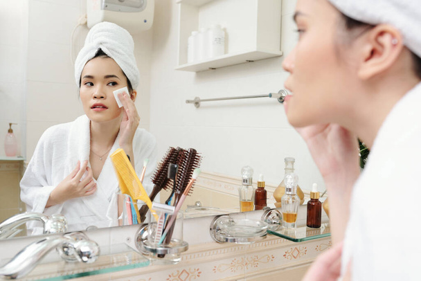 Giovane donna asiatica con morbida pelle liscia applicando toner con batuffolo di cotone quando si guarda finestra del bagno - Foto, immagini