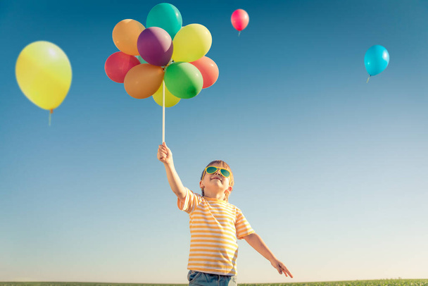 Happy child playing with bright multicolor balloons outdoor. Kid having fun in green spring field against blue sky background. Healthy and active lifestyle concept - Foto, Bild