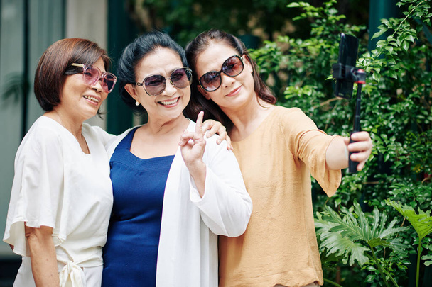 屋外に立つときにスマートフォンで自画撮りのためにポーズをとるサングラスで楽しいシニアアジアの女性 - 写真・画像