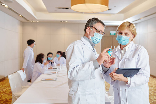 Mature experienced researcher in medical mask showing vial with newest version of vaccine to his coworker when their colleagues having discussion i background - Photo, Image