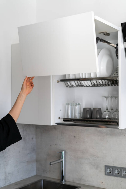 Vista vertical recortada de la mujer de pie en los armarios blancos de la cocina moderna, puerta abierta con costoso mecanismo de elevación - Foto, Imagen