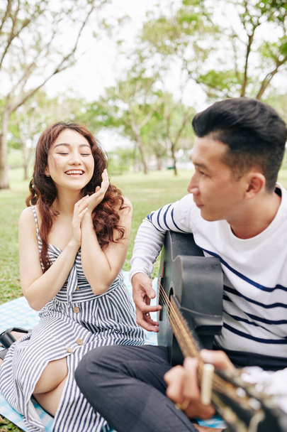 Happy excited young woman applauding her boyfriend singing and playing guitar when having romantic picnic in park - Фото, зображення