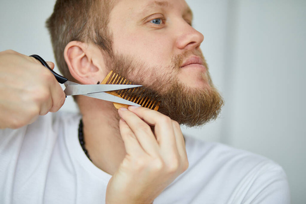 Красивий чоловік у білих футболках розрізає бороду, сам робить вуса ножицями вдома. Самодостатність під час карантину. Залишайтеся вдома
. - Фото, зображення