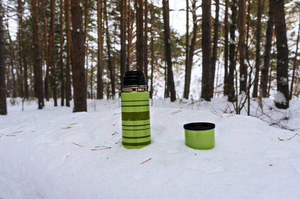 зелений смугастий термос, що стоїть на снігу на столі в лісі. Чайне пиття, зимовий похід
 - Фото, зображення