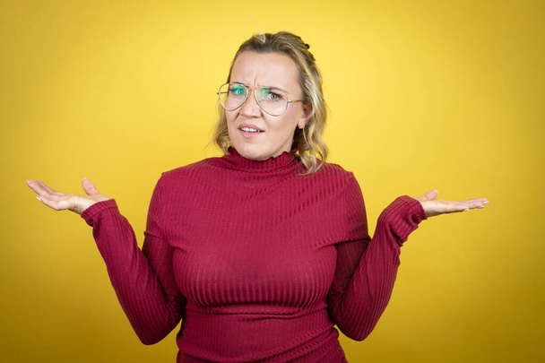 Young caucasian woman wearing casual red t-shirt over yellow background clueless and confused expression with arms and hands raised - Photo, Image