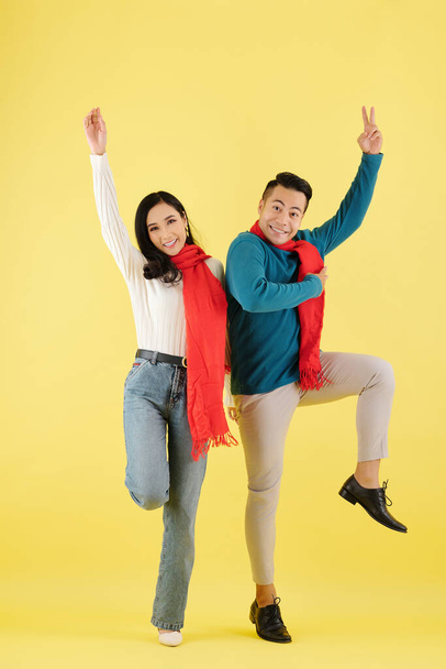 Excited happy young Asian couple in wool winter scarves dancing together against yellow background - Photo, Image