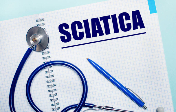 On a light blue background, an open notebook with the word SCIATICA, a blue pen and a stethoscope. View from above. Medical concept - Photo, image