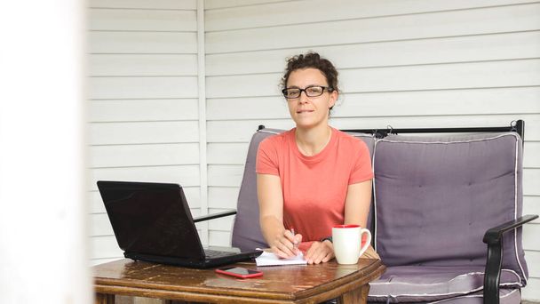 Mujer joven reflexiva en gafas de escribir notas, sentado en el sofá en la terraza del hogar con el ordenador portátil y una taza de café, mirando a la cámara. Aprender el idioma del estudiante, escuchar el curso en línea del webinar - Foto, Imagen