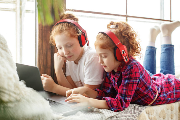 Two red-haired teenage girls in red headphones are merrily listening to music, next to a laptop, sitting on the window - Foto, afbeelding