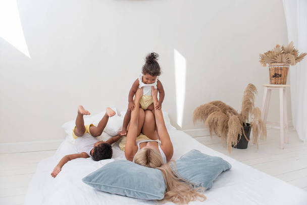 Young Caucasian blond mother plays, lies on white bed with African American daughter son. Happy multi ethnic family morning. Woman hugs, strokes children with tenderness. Minimalistic white interior - Valokuva, kuva