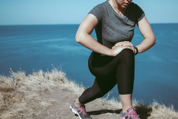 Sporty woman stretching legs and thigh muscles doing lunge exercise before running standing on cliff near blue sea background. Preparing muscles and ligaments before training workout. Copy space - Foto, afbeelding