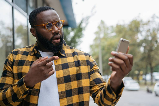 Emotional African American man shopping online, pointing finger on mobile phone screen. Influencer using smartphone, streaming video, communication with subscribers - Photo, Image