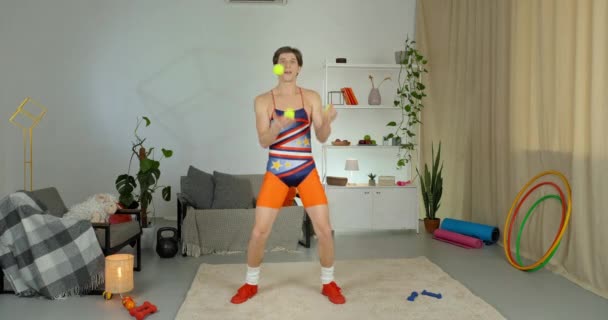 Blanke grappige lange en dunne mannelijke atleet in vintage trainingspak staat in de woonkamer rond sportuitrusting jongleren gele ballen gooit ze in de lucht met succes vangt, concept van evenwicht - Video