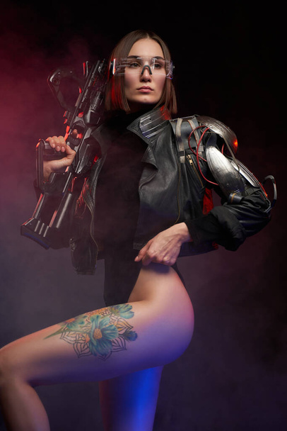 Sexy und schlanke Frau mit Implantat und Gewehr posiert vor dunklem Hintergrund - Foto, Bild