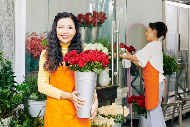 Portret młodej pięknej azjatyckiej kwiaciarni i z wiadrem czerwonych i białych róż stojących na wyświetlaczu kwiatowym, jej współpracownik sprayu kwiaty w tle - Zdjęcie, obraz