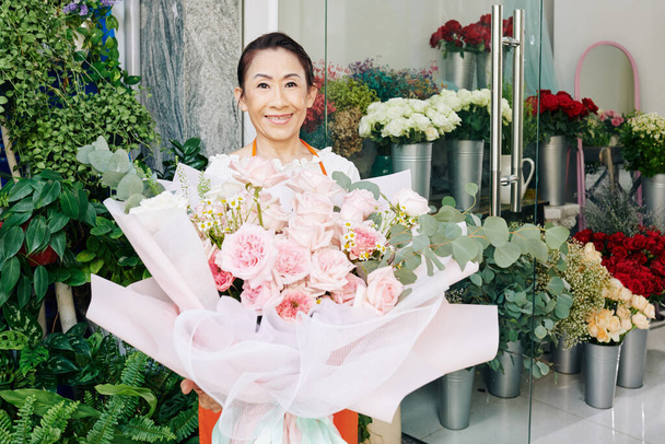 Портрет улыбающегося старшего владельца цветочного магазина, стоящего с большим светло-розовым букетом, который она создала для клиента - Фото, изображение