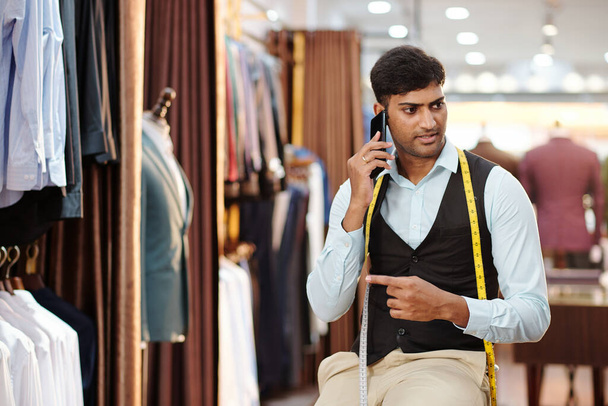 Portrait of serious young Indian tailor working in fashion atelier and talking on phone with client - Фото, изображение