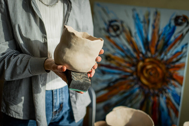 Красивая молодая женщина в творческой мастерской сидит за столом и вытирает глиняную чашу губкой. Формированный способ приготовления глиняных блюд. Ручная работа. Производство керамики - Фото, изображение