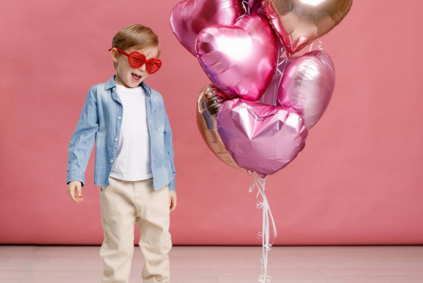 Boy dancing, smiling and having fun near  big branch of pink heart-shaped ballons. Isolated on pink background. - Photo, image