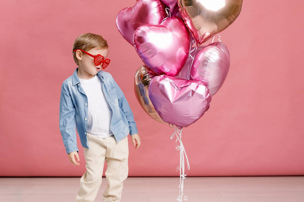 Boy dancing, smiling and having fun near  big branch of pink heart-shaped ballons. Isolated on pink background. - Fotografie, Obrázek