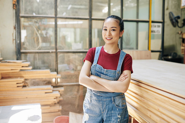 Portrait of pretty young Vietnamese carpenter in denim overall standing in carpentry workshop with arms folded - Valokuva, kuva