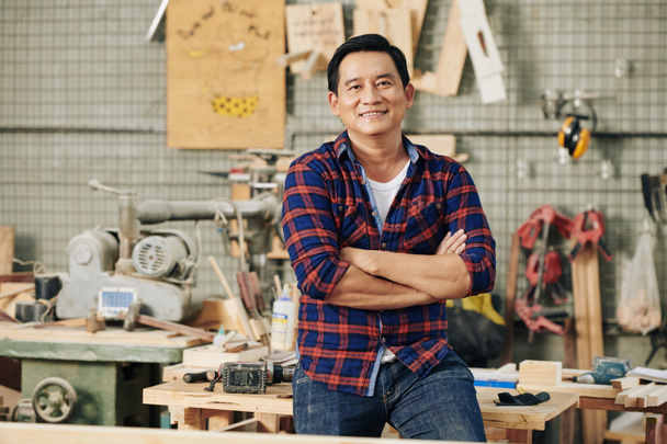 Carpinteiro confiante alegre em camisa xadrez em pé em sua oficina de carpintaria com os braços dobrados - Foto, Imagem