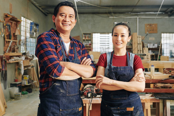 Веселые плотники, стоящие в мастерской в джинсовых фартуках, складывающие руки и смотрящие в камеру - Фото, изображение
