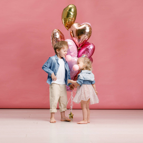Little boy and girl hugging, dancing, smiling and having fun near big branch of pink heart-shaped ballons. Valentins day concept. Isolated on pink background. - Photo, image