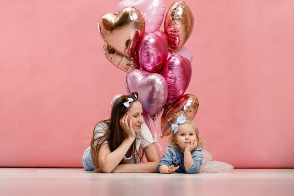 Young mother and her daughter dancing, smiling and having fun near  big branch of pink heart-shaped ballons. Isolated on pink background. - Fotografie, Obrázek