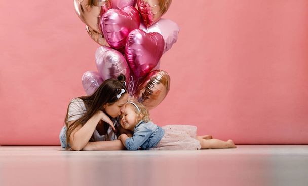 Joven madre y su hija bailando, sonriendo y divirtiéndose cerca de una gran rama de globos rosados en forma de corazón. Aislado sobre fondo rosa. - Foto, Imagen