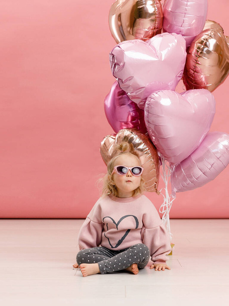 Cute little girl in glasses dancing, smiling and having fun near big branch of pink heart-shaped ballons. Isolated on pink background. - Photo, image