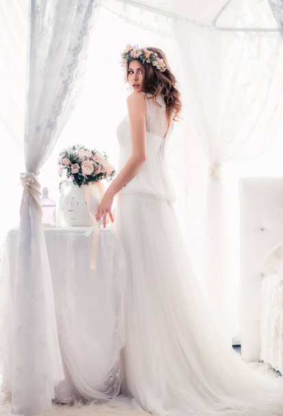 A bride in a wedding dress near the window. Beautiful tender morning with a decor of flowers - Фото, зображення