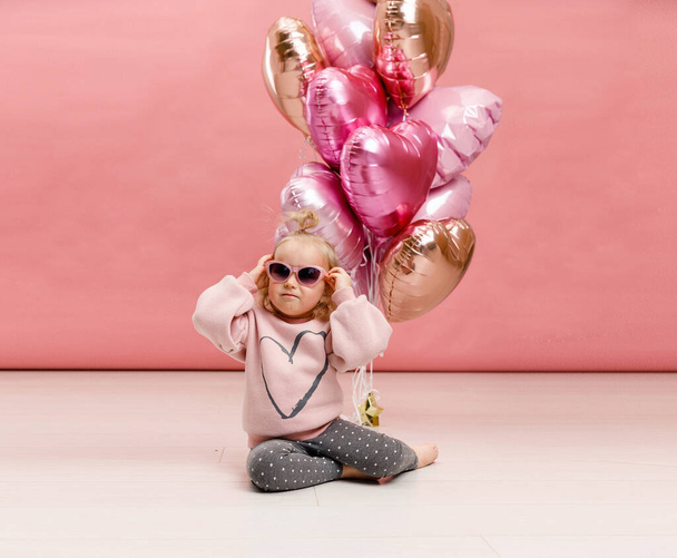 Cute little girl in glasses dancing, smiling and having fun near big branch of pink heart-shaped ballons. Isolated on pink background. - Fotoğraf, Görsel