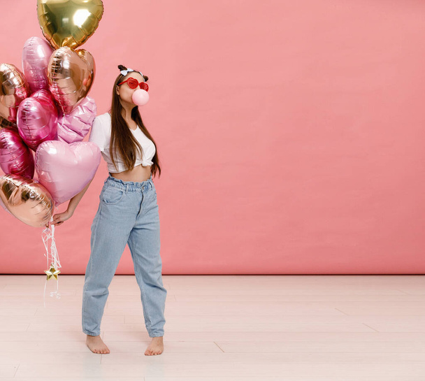 Young woman dancing, smiling and having fun with bubble gum near big branch of pink heart-shaped ballons. Isolated on pink background. - Foto, Imagem