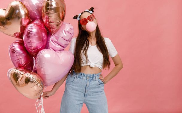 Young woman dancing, smiling and having fun with bubble gum near big branch of pink heart-shaped ballons. Isolated on pink background. - Foto, immagini