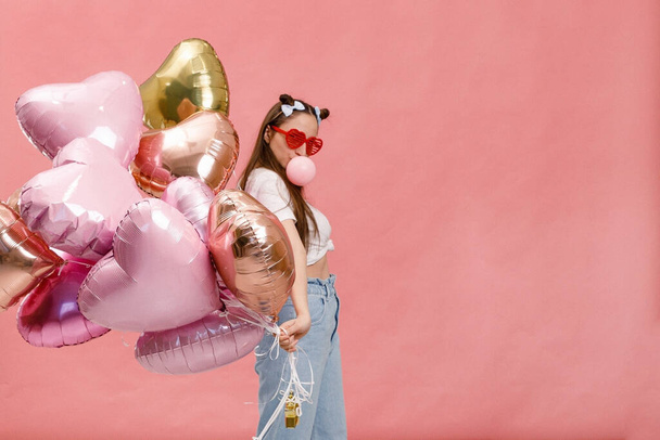Young woman dancing, smiling and having fun with bubble gum near big branch of pink heart-shaped ballons. Isolated on pink background. - Foto, Bild