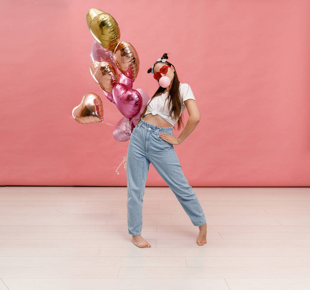 Mulher jovem dançando, sorrindo e se divertindo com chiclete perto de grande ramo de balões cor de rosa em forma de coração. Isolado sobre fundo rosa. - Foto, Imagem
