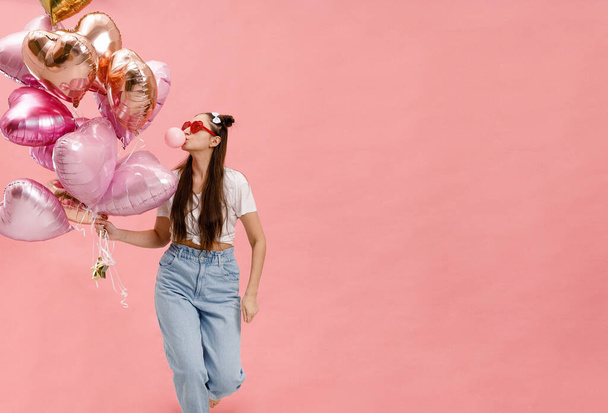 Young woman dancing, smiling and having fun with bubble gum near big branch of pink heart-shaped ballons. Isolated on pink background. - Fotografie, Obrázek