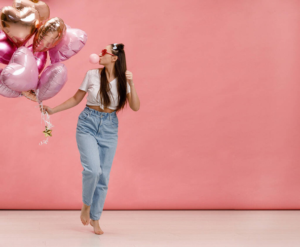 Young woman dancing, smiling and having fun with bubble gum near big branch of pink heart-shaped ballons. Isolated on pink background. - Foto, imagen