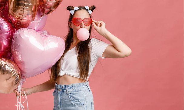 Young woman dancing, smiling and having fun with bubble gum near big branch of pink heart-shaped ballons. Isolated on pink background. - Photo, Image