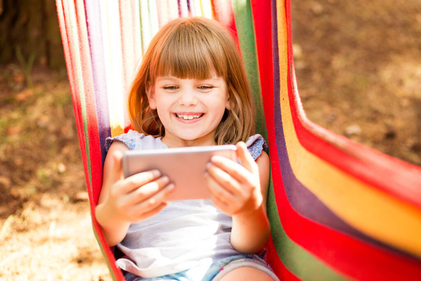Usmívající se holčička nahrává blog o životním stylu, mluví s kamerou mobilního telefonu venku, sedí na houpací síti. Šťastný mladý ovlivňovač natáčí vlog pro kanál. Koncepce blogování.  - Fotografie, Obrázek