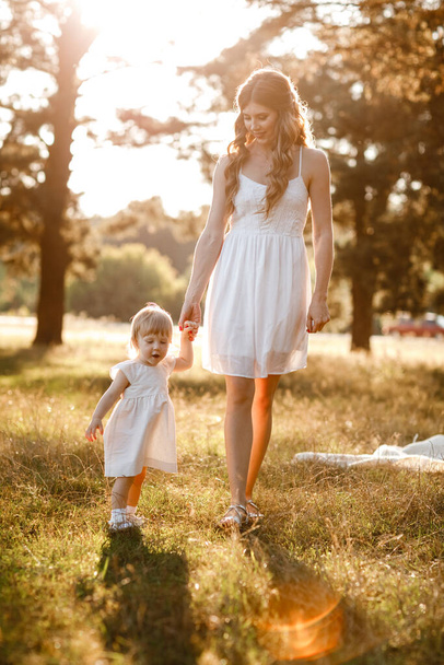 Η νεαρή όμορφη μαμά περπατάει με την κόρη της στο ηλιόλουστο πάρκο. Η έννοια των καλοκαιρινών διακοπών σε εξωτερικούς χώρους. Της μητέρας, την ημέρα του μωρού. Περνάμε χρόνο μαζί. Ηλιακό φως - Φωτογραφία, εικόνα