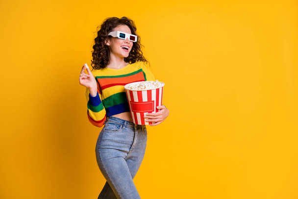 Photo of cheerful funky young brunette hair lady stand eat popcorn watch film wear spectacles sweater jeans isolated on yellow background - Fotó, kép