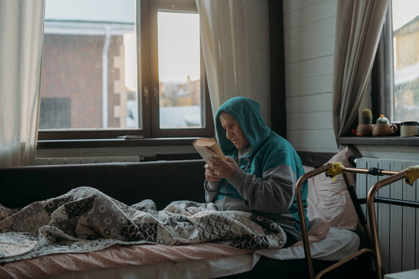 Η γιαγιά κάθεται στον καναπέ και διαβάζει ένα βιβλίο. Μια ηλικιωμένη γυναίκα, συνταξιούχος, αναπαύεται στο σαλόνι κατά τη διάρκεια της ημέρας. Περιπατητές δίπλα στο κρεβάτι - Φωτογραφία, εικόνα